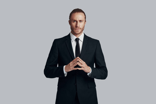 Energy inside him. Handsome young man in full suit looking at camera and keeping hands clasped while standing against grey background - Photo, Image