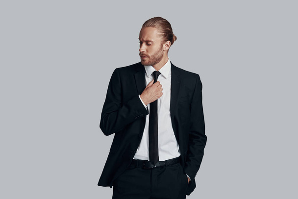 Young and successful. Handsome young man in full suit adjusting his necktie while standing against grey background - Фото, зображення