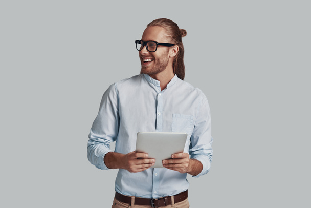 Positiv charmant. schöner junger Mann, der mit Laptop arbeitet und vor grauem Hintergrund lächelt - Foto, Bild