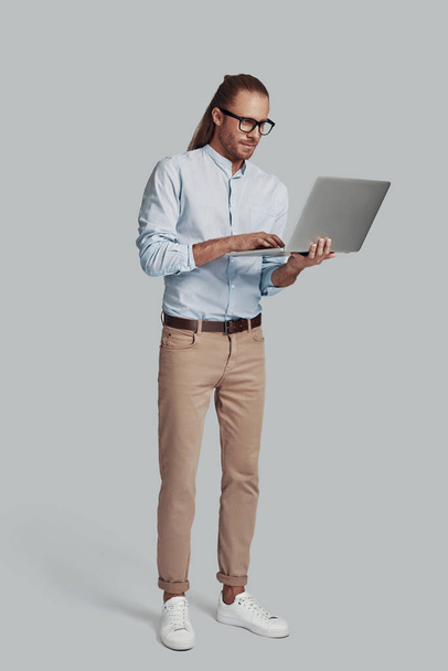 Itsevarma liikemies. Täysi pituus komea nuori mies työskentelee kannettavan tietokoneen kun seisoo harmaata taustaa vasten
 - Valokuva, kuva