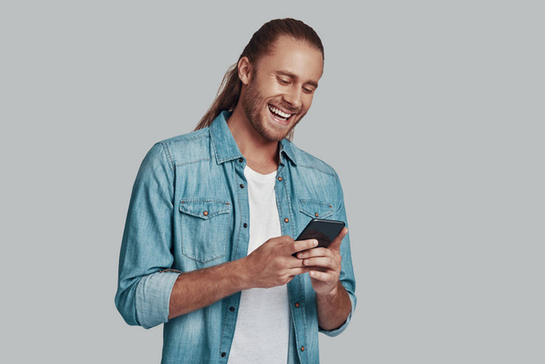 Κοινωνική συγκέντρωση. Όμορφος νέος άντρας που χρησιμοποιεί έξυπνο τηλέφωνο και χαμογελά ενώ στέκεται ενάντια στο γκρίζο φόντο - Φωτογραφία, εικόνα