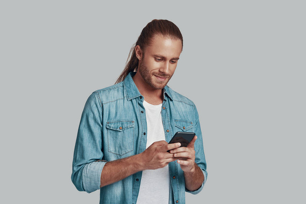 Społecznościowych. Przystojny młody człowiek przy pomocy silny telefon i uśmiechnięty chwila stojący wobec szary tło - Zdjęcie, obraz