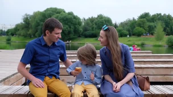 Happy Family Concept: mother, father and little boy sitting by the river and eating croissant - 映像、動画