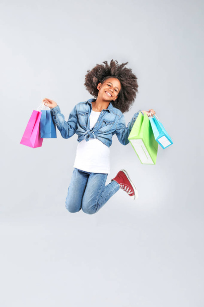 Jumping African-American girl in jeans clothes and with shopping bags on grey background - Photo, Image