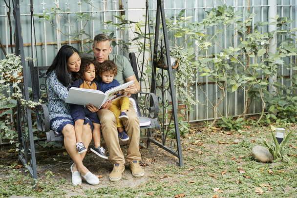 Счастливая семья смешанных рас сидит на качелях и читает сказки вместе
 - Фото, изображение