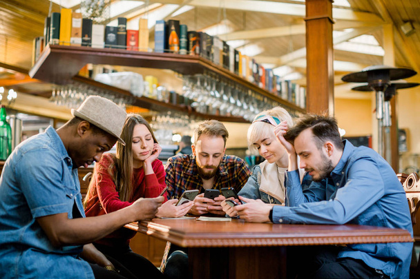 Αντίληψη του ψηφιακού tablet για την τεχνολογία επικοινωνίας των ατόμων. Ομάδα των πέντε πολυεθνικικών μαθητών που κάθονται σε ένα καφέ μπαρ κοιτάζοντας smartphones-νέοι χαρούμενοι φίλοι διασκεδάζουν - Φωτογραφία, εικόνα