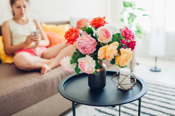 Jovem mulher usando smartphone sentado no sofá em casa. Sala de estar decorada wih buquê de rosas
 - Foto, Imagem
