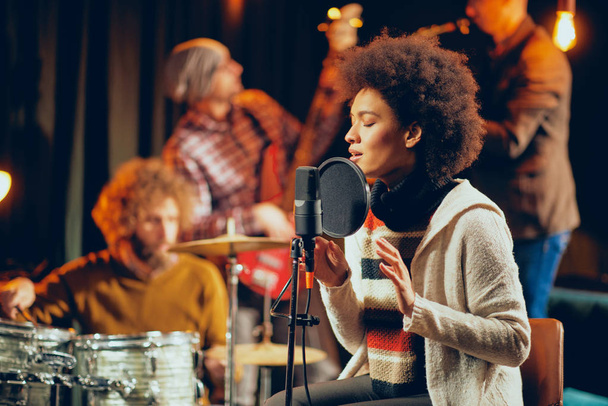 Mixed race woman singing. In background band playing instruments. Home studio interior. - Photo, Image