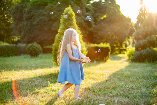 Child whirling, dancing plays on the meadow. Girl having fun with bubbles. Cute little longhair blonde girl dancing with soap bubbles at sunset park - Photo, image