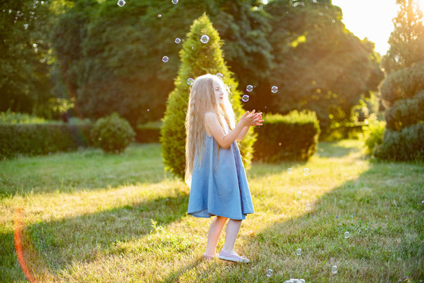 menina de pele bonita com longos cachos loiros em vestido branco brincando com bolhas de sabão no prado de verão no dia ensolarado brilhante
 - Foto, Imagem
