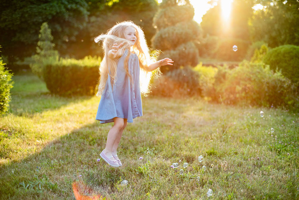 Un niño girando, bailando en el prado. Chica divirtiéndose con burbujas. Linda chica rubia de pelo largo bailando con burbujas de jabón en el parque del atardecer - Foto, imagen