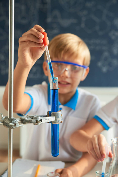 Schoolboy adicionando reagente azul no tubo de ensaio durante a realização do experimento
 - Foto, Imagem