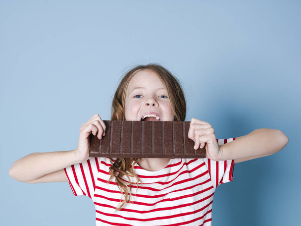 pretty blonde girl with giant bar of chocolate posing in front of blue background - Φωτογραφία, εικόνα