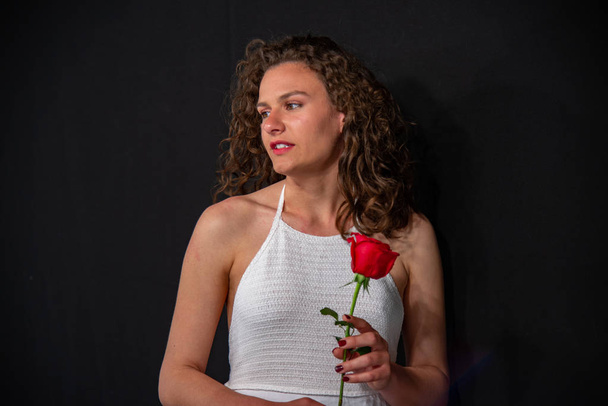 Half-length portrait of a beautiful sexy girl in a white sleeveless tunic. She is holding a red rose flower. On a uniform black background, copy space. - 写真・画像