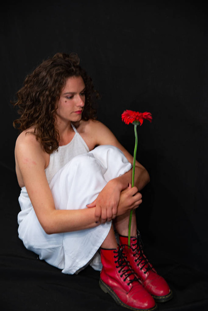 Gyönyörű szexi lány ülve, fehér ujjatlan tunikus, piros cipők csizma. Ő kezében egy piros Gerbera virág. Egy egységes fekete háttér, a másolás Space. - Fotó, kép