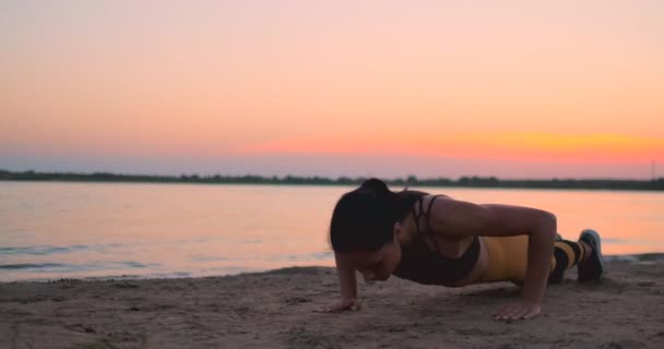 Sporty girl doing push ups on beach at sunrise. Young fit tanned smiling girl in sport top and leggings doing push ups on beach at sunset. - Footage, Video