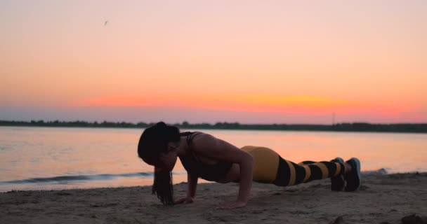 Sporty girl doing push ups on beach at sunrise. Young fit tanned smiling girl in sport top and leggings doing push ups on beach at sunset. - Footage, Video