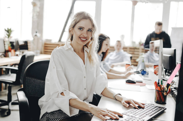 Young beautiful woman dressed in white shirt is working at the laptop sitting at the desk in a light modern open space office - Foto, Bild