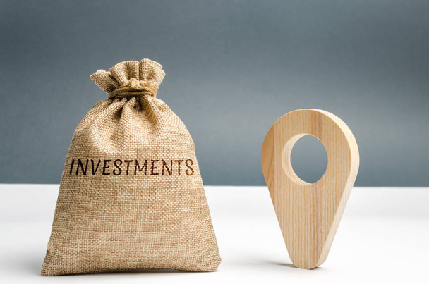 Money bag with the word Investments and a geolocation marker. Point and direct investments in a region or country. Construction of new factories and infrastructure. Economic attractiveness - Fotó, kép