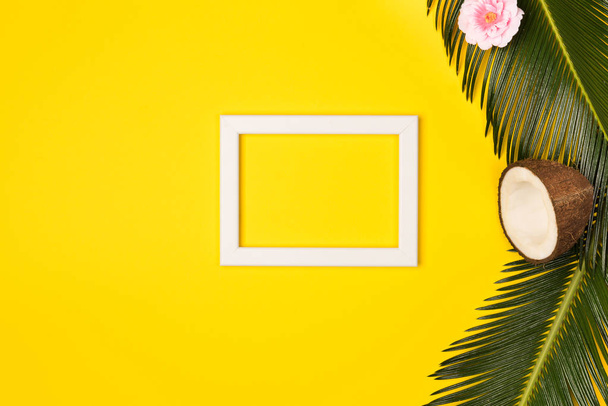 Stylish summer composition with photo frame, green leaves, flower and coconut on a yellow background. Artwork mockup with copy space - Foto, imagen