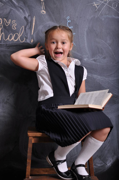 Retrato de belo sorridente jovem do primeiro ano sentado com livro sobre fundo quadro-negro. conceito de escola e educação
 - Foto, Imagem