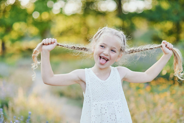 Funny girl holding hair plaits. Small cute girl with long blonde hair showing tongue outdoor. Kid expressing emotions. April fool. - Fotó, kép