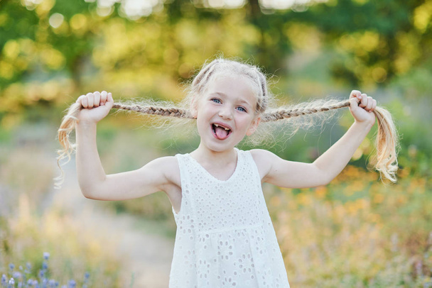 Забавна дівчина тримає зачіски. Маленька мила дівчинка з довгим світлим волоссям, що показує язик на відкритому повітрі. Малюк висловлює емоції. квітня дурень
. - Фото, зображення