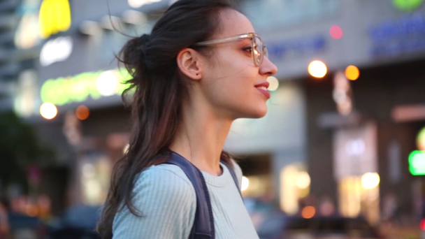 ύπαιθρο αστικό πορτραίτο ενός νεαρού ενήλικα όμορφη γυναίκα σε γυαλιά, που ποζάρει σε εξωτερικούς χώρους στο δρόμο για τη νυχτερινή πόλη φώτα φόντο. σύγχρονη νεαρή γυναίκα περπάτημα πόλη το καλοκαίρι το βράδυ - Πλάνα, βίντεο