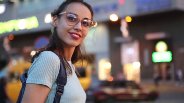 ύπαιθρο αστικό πορτραίτο ενός νεαρού ενήλικα όμορφη γυναίκα σε γυαλιά, που ποζάρει σε εξωτερικούς χώρους στο δρόμο για τη νυχτερινή πόλη φώτα φόντο. σύγχρονη νεαρή γυναίκα περπάτημα πόλη το καλοκαίρι το βράδυ - Πλάνα, βίντεο