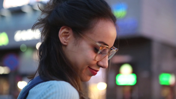 outdoors urban portrait of a young adult beautiful woman in glasses, posing outdoors on the street on night city lights background. modern young woman walking city at summer evening - Felvétel, videó