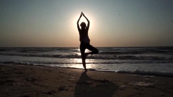 Κορίτσι στέκεται σε μια πόζα γιόγκα, απέναντι από τον ήλιο στο ηλιοβασίλεμα, στην παραλία, στη θάλασσα - Πλάνα, βίντεο