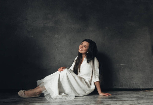 Young beautiful woman sitting on the floor at home. Smiling girl in white dress near black wall.  - Fotoğraf, Görsel