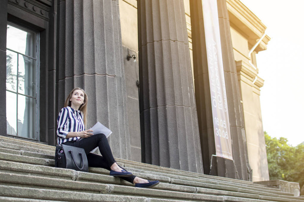όμορφος μαθητής κάθεται στα σκαλιά κοντά στο κολλέγιο με ένα σακίδιο, κρατώντας χαρτιά στα χέρια της και κοιτάζει προς τα πάνω, μαθαίνει μαθήματα στο διάλειμμα - Φωτογραφία, εικόνα