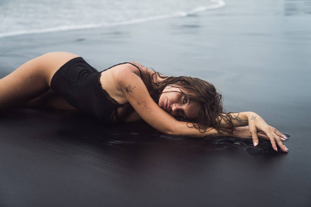 Sexy tanned girl with wet hair in swimwear lies on deserted black sandy beach - Foto, Imagem