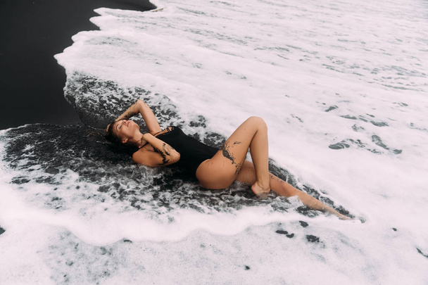 Sensual sexy girl in swimsuit lying on black sand beach in ocean waves - Фото, изображение