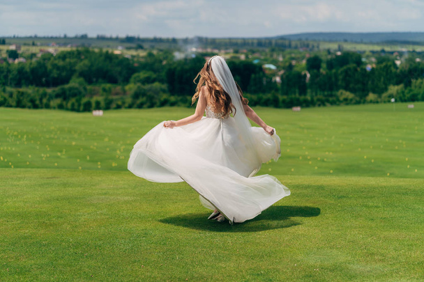 Backside view, bride runs in white wedding dress with veil on green field - Foto, Imagen