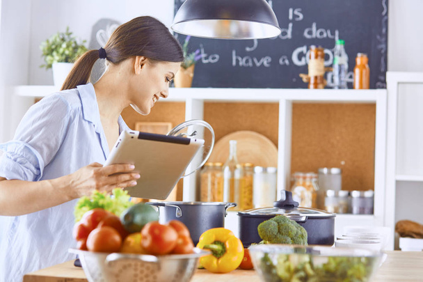 Obrázek Mladá hezká holka stojí v kuchyni a vaření potravin. Při pohledu na tabletový počítač - Fotografie, Obrázek