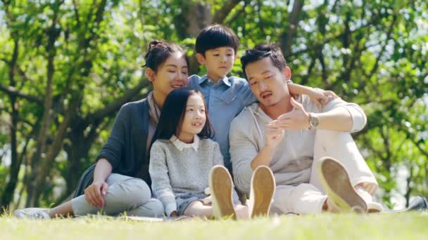 Azjatycka rodzina z dwójką dzieci siedzi na trawie na zewnątrz w parku rozmawiając na czacie - Materiał filmowy, wideo