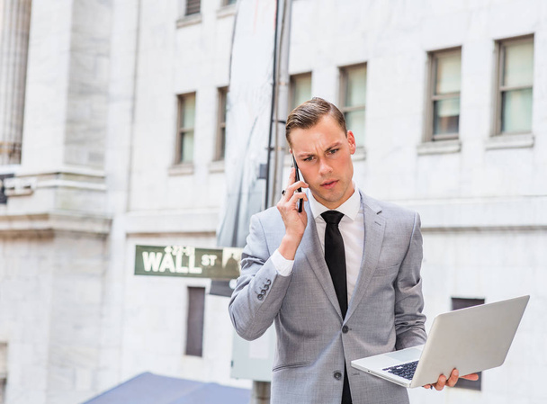 ニューヨークで働いている若いビジネスマンがグレーのブレザー、白いシャツ、黒いネクタイを着て、ウォール街のサインで外のオフィスに立って、ラップトップコンピュータで作業し、携帯電話で話しています - 写真・画像