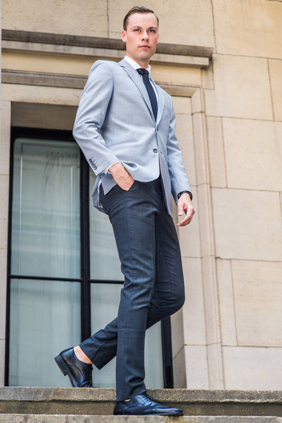 Young Businessman Street Fashion in New York City. Young Man wearing gray blazer, white shirt, black tie, black pants, leather shoes, walking down stairs by window outside office building, looking - Foto, afbeelding