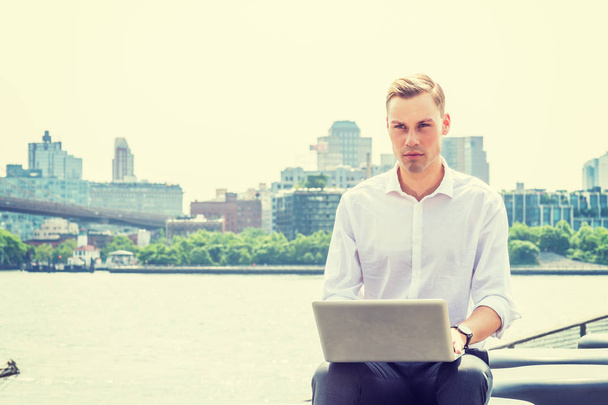 junger europäischer Geschäftsmann auf Reisen, arbeitet in New York City, trägt weißes Langarmshirt, sitzt am East River, arbeitet am Laptop, schaut, denkt. Brooklyn-Gebäude im Hintergrund - Foto, Bild