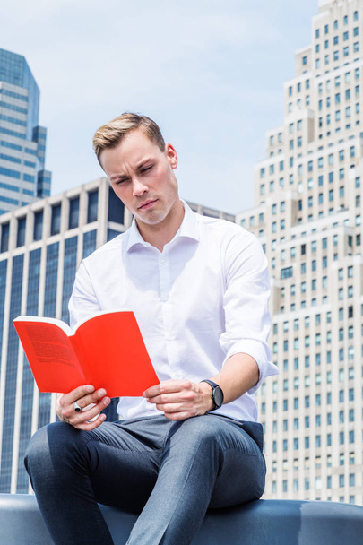 Ich lese gerne. junger gutaussehender europäischer Mann mit weißem Langarmshirt, schwarzer Hose, Armbanduhr, sitzt im Geschäftsviertel mit hohen Gebäuden in New York City, unter der Sonne, liest rotes Buch - Foto, Bild