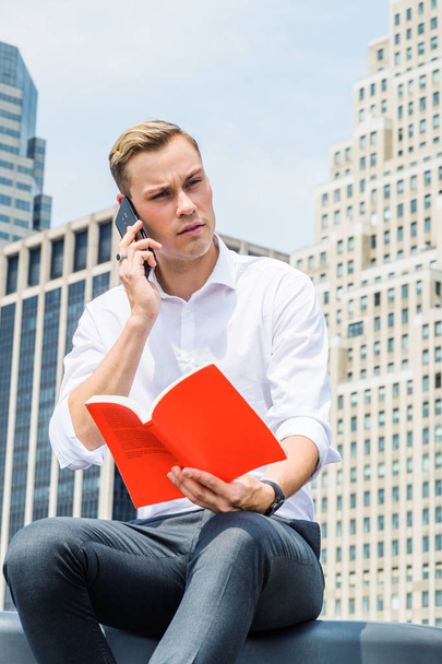 Young Handsome European Man wearing white long sleeve shirt, black pants, sitting in business district with high buildings in New York City, under sun, reading red book, talking on cell phone - Photo, Image