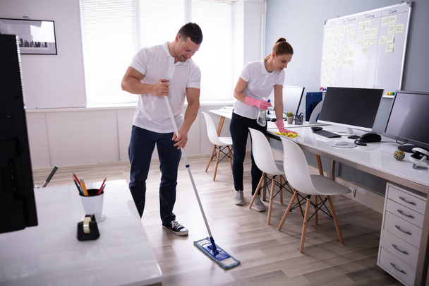 Two Smiling Young Janitor Cleaning The Desk And Mopping Floor In The Office - Zdjęcie, obraz
