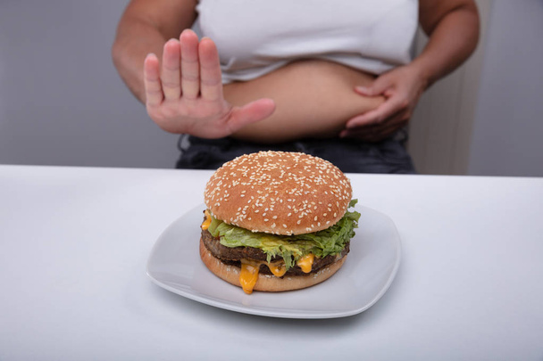 Mid Section Of Overweight Woman Showing No Gesture To Hamburger On Plate Over The Table - Foto, imagen