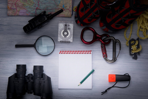 on a gray background, is a set of traveler comfortable shoes, rope, carabiners, magnifying glass, binoculars, compass, map, walkie-talkie, flashlight, in the center of a white notebook for writing with a pencil - Photo, image