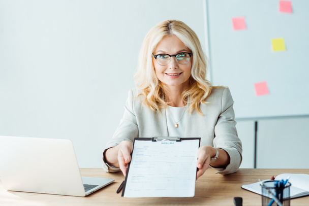 blonde cheerful woman in glasses holding clipboard with resume while sitting near desk  - Фото, зображення