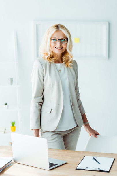 cheerful recruiter in glasses standing near chair in office - Photo, Image