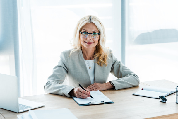 cheerful blonde woman in glasses looking at clipboard near laptop  - Photo, Image