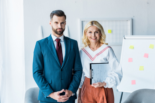 handsome recruiter with clenched hands and blonde woman with clipboard standing in office  - Фото, изображение